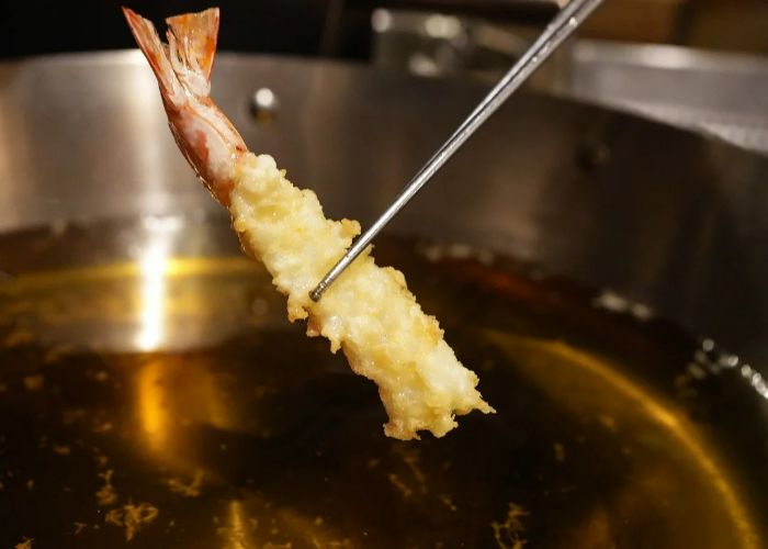 A crispy prawn tempura being lift with chopsticks from a vat of oil.